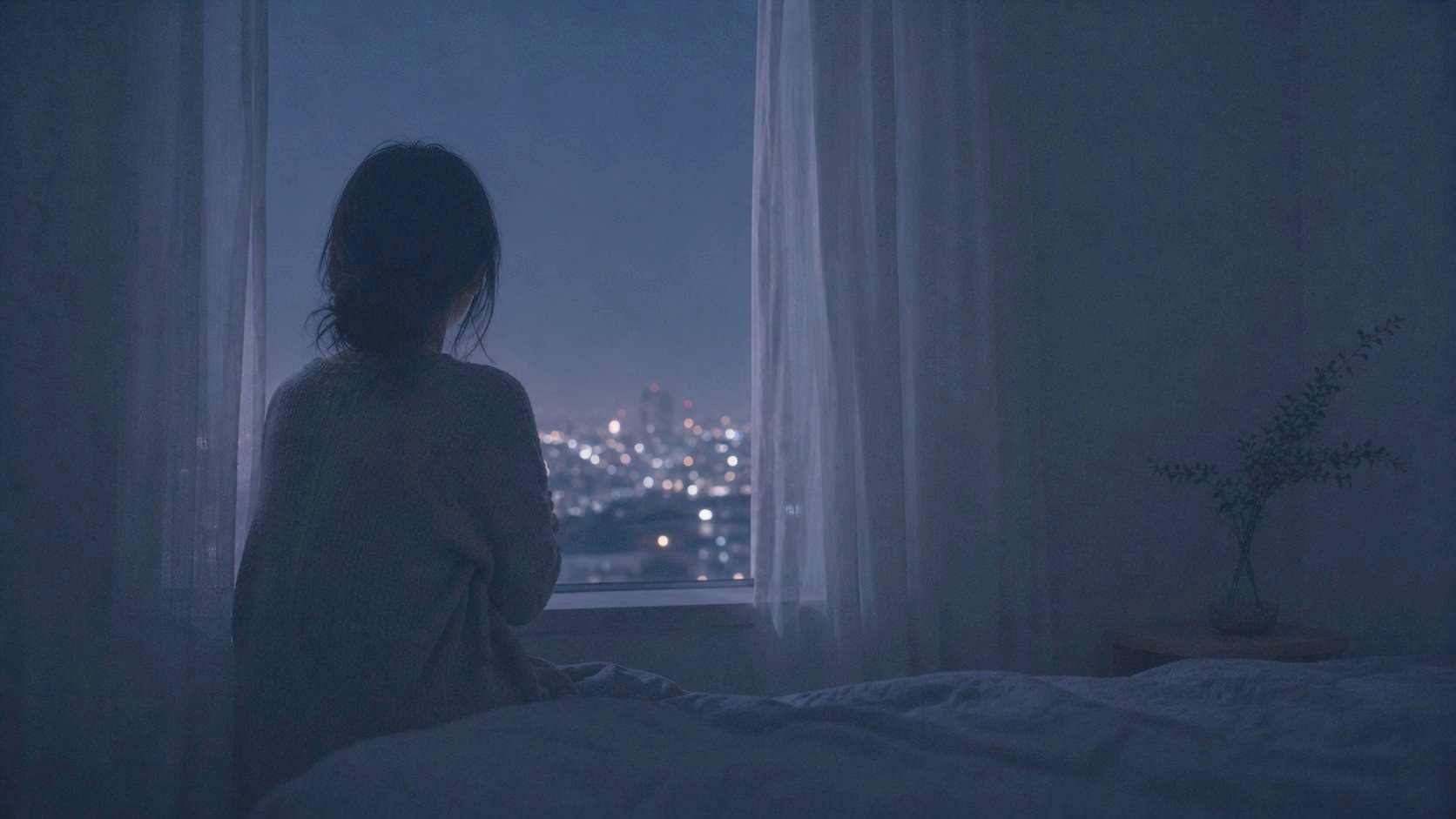 夜、ベッドに腰掛けて窓の外の夜景を見る後ろ姿