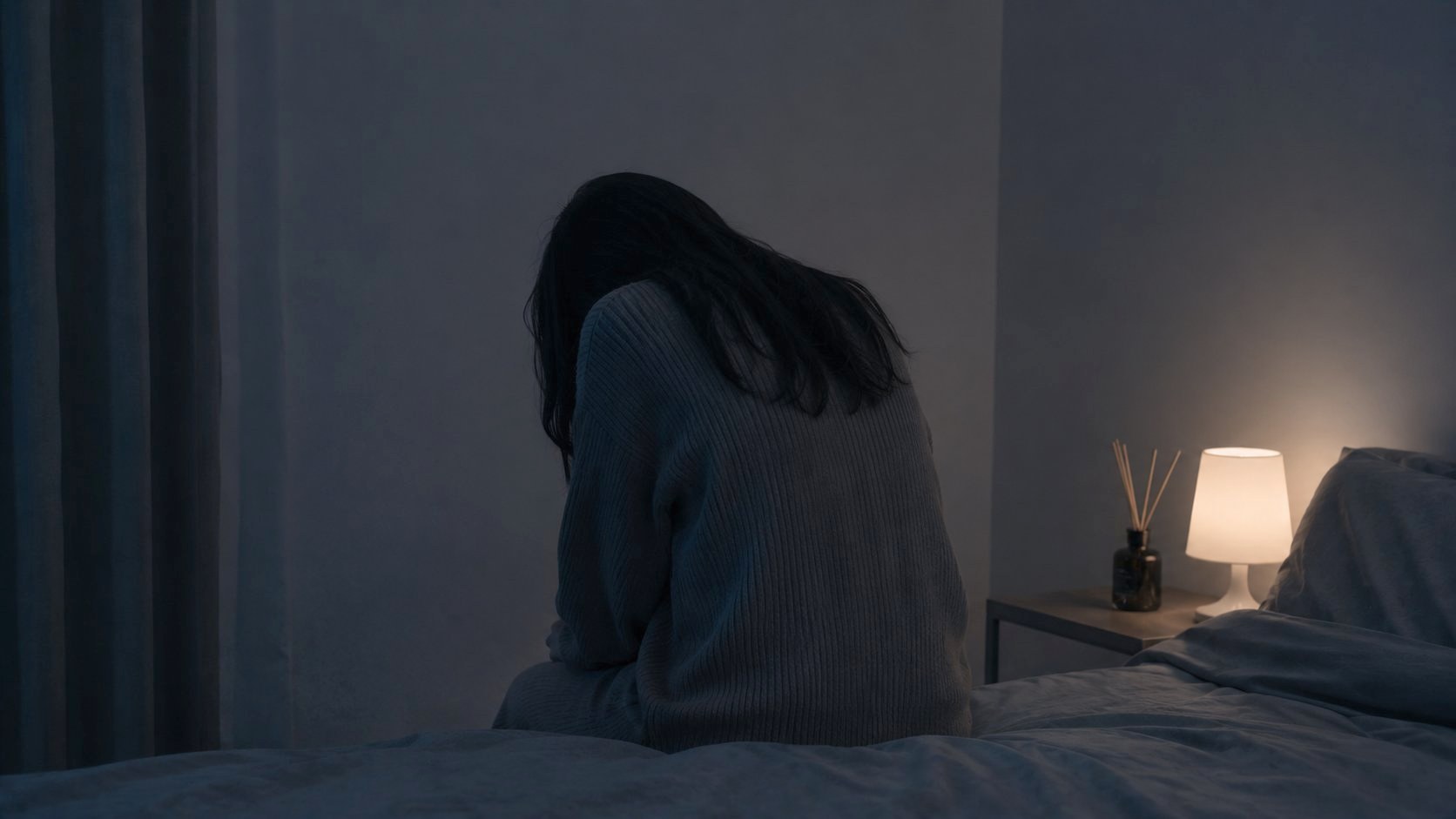 夜、ベッドの縁に腰掛けて深くうつむく後ろ姿